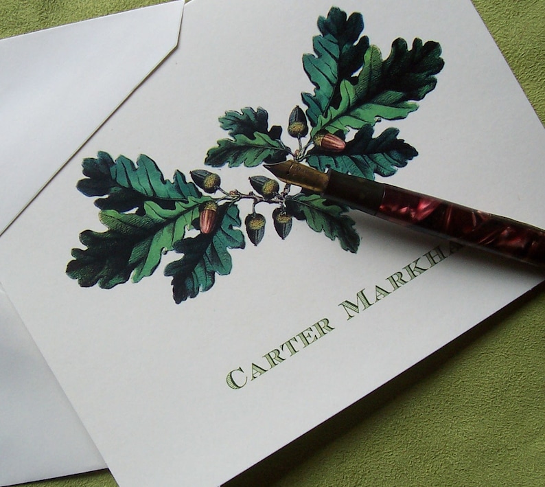 K&ouml;nnte beinhalten: Eine wei&szlig;e Karte mit einem gr&uuml;nen Eichenblatt-Design und Eicheln. Der Text "CARTER MARKHA" ist in Gr&uuml;n auf die Karte gedruckt. Ein rot-goldener Stift liegt auf der Karte.