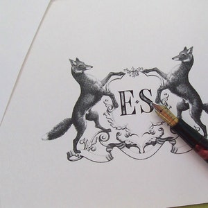 May include: A black and white illustration of two foxes flanking a crest with the initials "ES" in the centre. A vintage-style fountain pen is lying on the paper.