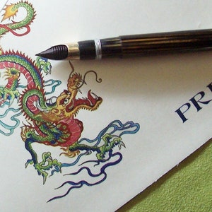 May include: A green and red dragon with blue and white clouds is drawn on a white paper. A black and gold fountain pen is placed on the paper above the dragon. The text "PRB" is written in blue ink on the paper.