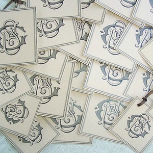 May include: A stack of blank, cream-colored gift tags with a black and gray monogram design. The tags have a string attached to the top.
