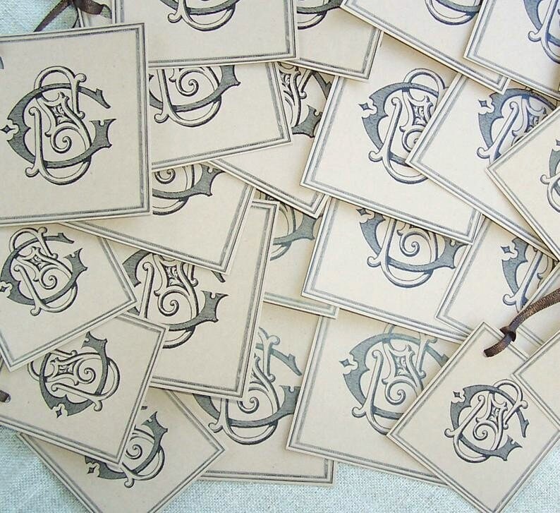 May include: A stack of blank, cream-colored gift tags with a black and gray monogram design. The tags have a string attached to the top.