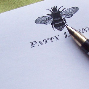 May include: A white envelope with the name "PATTY L" printed in black. A black and white bee illustration is above the name. A black pen with a gold tip is lying on the envelope.