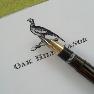 May include: A black and white illustration of a pheasant on a white card with a black fountain pen resting on the card. The card reads "Oak Hill Manor".