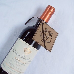May include: A bottle of Chateau Du Buisin Bordeaux wine with a brown paper gift tag. The tag has a monogram with the letters "Y" and "C" in a decorative font.