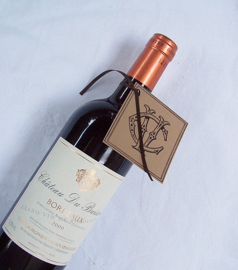May include: A bottle of Chateau Du Buisin Bordeaux wine with a brown paper gift tag. The tag has a monogram with the letters "Y" and "C" in a decorative font.