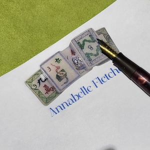 May include: A close-up of a stack of four white tiles with Chinese characters and green and red accents. The tiles are on a white surface with the name "Annabelle Fletcher" written in blue ink. A brown and gold fountain pen is lying on the surface.