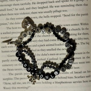 May include: A multi-strand beaded bracelet featuring black and white letter beads spelling out the phrase MY BOY ONLY BREAKS HIS TOYS. The bracelet includes small faceted black beads, silver-toned metal charms, and a small silver-toned lightning bolt charm.