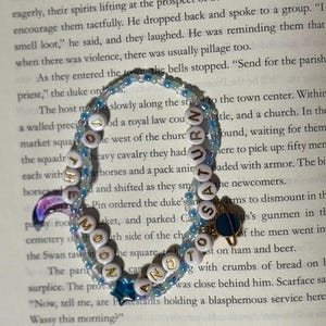 May include: A double-strand beaded bracelet featuring light blue and clear beads, white letter beads spelling out THE MOON AND TO SATURN, a purple crescent moon charm, a blue star charm, and a small gold-toned planet charm with a blue ring.