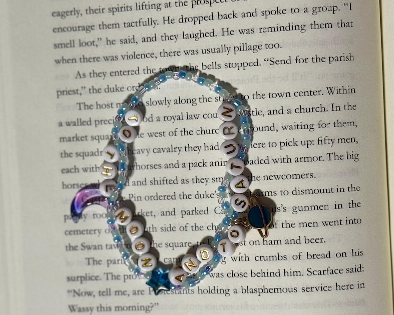 May include: A double-strand beaded bracelet featuring light blue and clear beads, white letter beads spelling out THE MOON AND TO SATURN, a purple crescent moon charm, a blue star charm, and a small gold-toned planet charm with a blue ring.