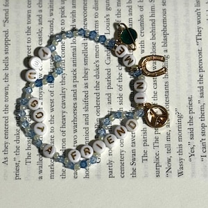May include: A beaded bracelet featuring light blue and clear crystal beads interspersed with white alphabet beads spelling out "YOU GOT A FRIEND IN ME". The bracelet includes gold-toned charms shaped like a planet, a horseshoe, and a peace sign.