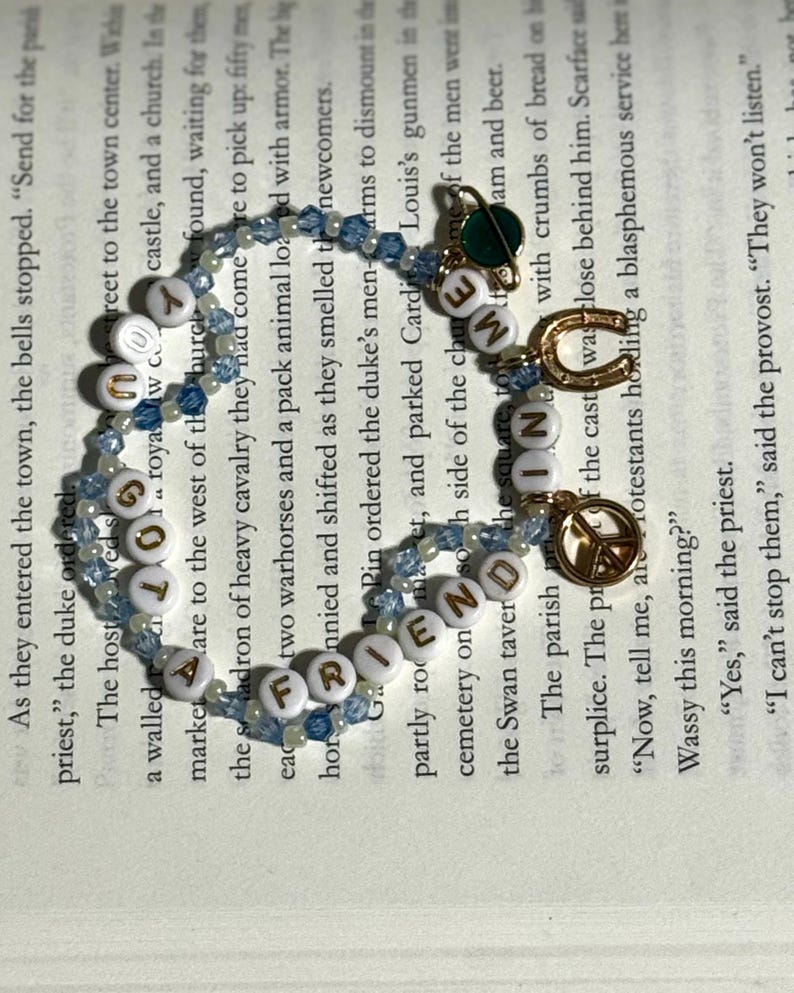 May include: A beaded bracelet featuring light blue and clear crystal beads interspersed with white alphabet beads spelling out "YOU GOT A FRIEND IN ME". The bracelet includes gold-toned charms shaped like a planet, a horseshoe, and a peace sign.