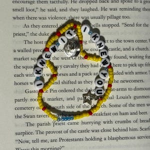 May include: A beaded bracelet featuring yellow, red, and blue seed beads with white letter beads spelling out I KNEW IT I KNEW YOU. The bracelet includes a silver-toned circular charm and a small silver-toned star charm attached to the strand.