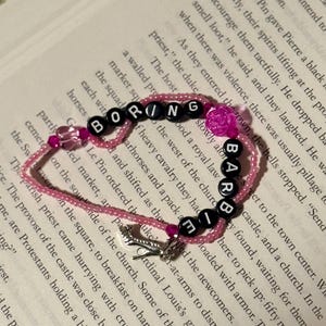 May include: A beaded bracelet featuring pink translucent beads, black letter beads spelling out BORING BARBIE, a translucent pink rose-shaped bead, a small pink heart-shaped bead, and a silver-toned high-heeled shoe charm dangling from the strand.