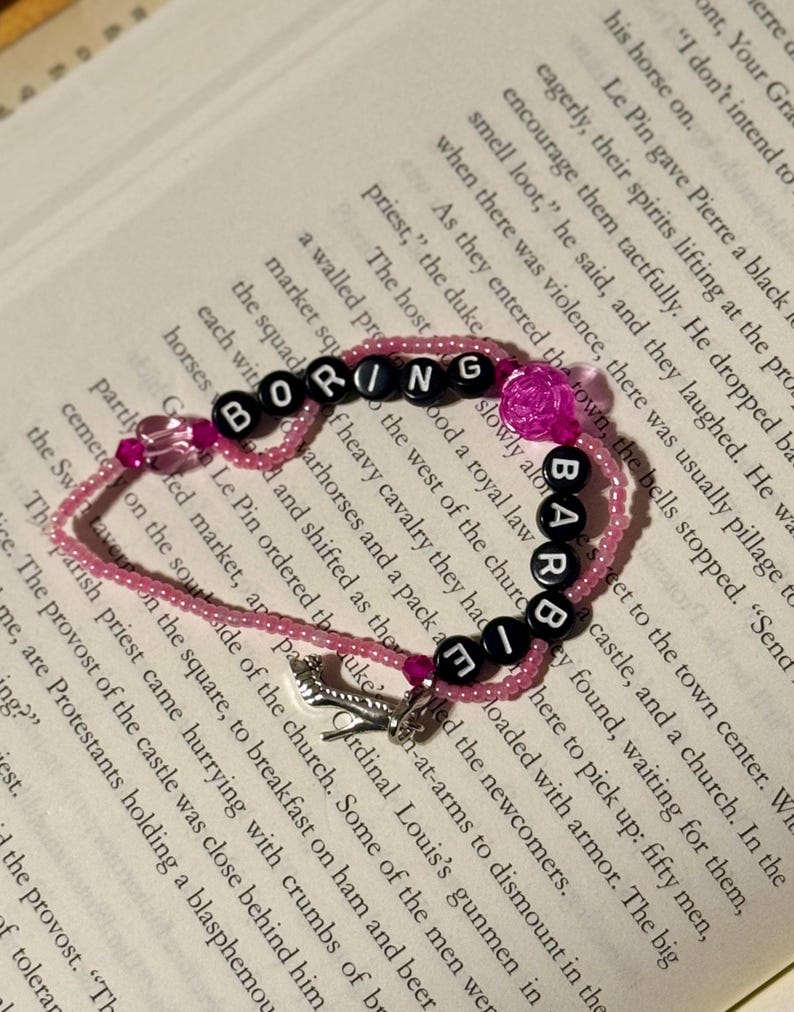 May include: A beaded bracelet featuring pink translucent beads, black letter beads spelling out BORING BARBIE, a translucent pink rose-shaped bead, a small pink heart-shaped bead, and a silver-toned high-heeled shoe charm dangling from the strand.
