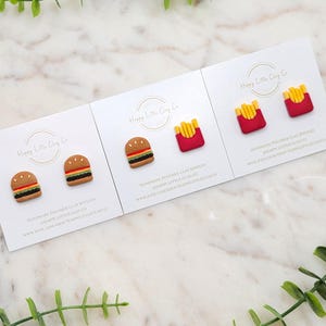May include: Polymer clay earrings featuring miniature food designs. The image displays three sets of earrings: burgers, and french fries. Each set is mounted on a white card with the text "Happy Little Clay Co" and the shop's website.