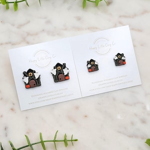 May include: Two pairs of Halloween-themed polymer clay earrings. Each earring features a miniature haunted house with a black roof, brown door, and orange accents. White ghosts and a jack-o'-lantern are also present. The earrings are displayed on white cards with the text "Happy Little Clay Co."