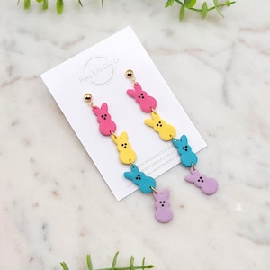 Puede incluir: Un par de pendientes colgantes con cuatro formas de conejito en colores pastel. Los conejitos son rosa, amarillo, azul y morado. Los pendientes están hechos de arcilla polimérica y tienen ganchos dorados.