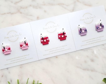 May include: Polymer clay earrings in pink, red, and purple, featuring a Snoopy-inspired design. The earrings are displayed on white cards with the Happy Little Clay Co. logo and website address. The background includes greenery.