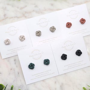 Puede incluir: Pendientes de botón en varios colores, incluyendo beige, gris, óxido, verde oscuro y negro, exhibidos en tarjetas blancas. Cada par presenta un diseño de nudo. Las tarjetas tienen el texto "Happy Little Clay Co" y "Handmade Polymer Clay Jewelry".