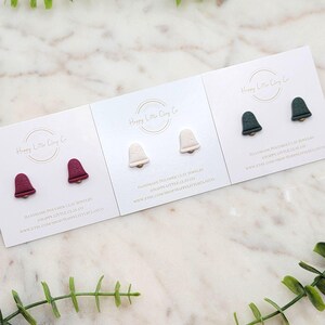 May include: Three pairs of bell-shaped polymer clay earrings in burgundy, cream, and dark green, each with a gold-colored post. The earrings are displayed on white cards with the text "Happy Little Clay Co" and website information.