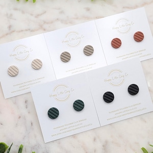 May include: Five pairs of handmade polymer clay stud earrings in beige, taupe, rust, dark green, and black. Each pair has a circular design with horizontal lines. The earrings are displayed on white cards with the text "Happy Little Clay Co."