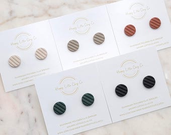 May include: Five pairs of handmade polymer clay stud earrings in various colors: beige, taupe, rust, dark green, and black. Each pair features a circular design with horizontal lines. The earrings are displayed on white cards with the text "Happy Little Clay Co."