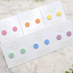 May include: Six pairs of handmade polymer clay earrings on white cards. The earrings are in various colors, including pink, orange, yellow, green, blue, and purple. Each pair has a striped pattern. The cards have the text "Happy Little Clay Co." and a website address.