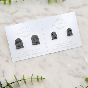 May include: Two pairs of gray polymer clay earrings shaped like tombstones, each with the letters "RIP" engraved on them. The earrings are displayed on white cards with the text "Happy Little Clay Co" and website information. Green foliage surrounds the cards.