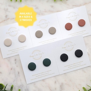 May include: Four sets of handmade polymer clay stud earrings in beige, taupe, terracotta, dark green, and black. Each pair is presented on a white card with the Happy Little Clay Co. logo. The text "AVAILABLE IN 4 SIZES & 2 FINISHES" is also visible.