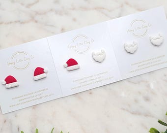 Puede incluir: Tres juegos de pendientes navideños de arcilla polimérica sobre tarjetas blancas. Un juego presenta gorros de Papá Noel con detalles rojos y blancos. Otro juego muestra barbas blancas de Papá Noel. Las tarjetas tienen el texto "Happy Little Clay Co."