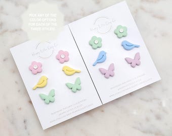Puede incluir: Dos tarjetas con una variedad de pendientes de arcilla polimérica de colores. Los pendientes tienen forma de flores, mariposas y pájaros. Las tarjetas tienen el texto "Happy Little Clay Co" y "Pendientes de arcilla polimérica hechos a mano".