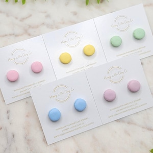 Könnte beinhalten: Vier Paar handgefertigte Polymer Clay Ohrringe in Form von Macarons. Die Farben sind rosa, gelb, grün und blau. Die Ohrringe werden auf weißen Karten mit dem Text "Happy Little Clay Co." und Informationen über die Ohrringe präsentiert.