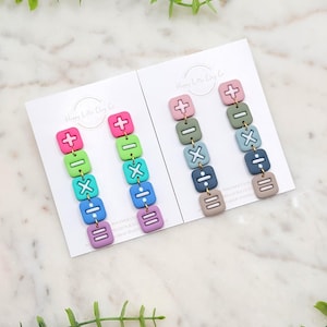 Puede incluir: Dos pares de pendientes de arcilla polimérica coloridos. Un par presenta un vibrante degradado de símbolos de calculadora rosas, verdes, azules y morados. El otro par tiene una paleta apagada de símbolos de calculadora rosas, verdes, azules y taupe. Cada pendiente tiene un signo más, menos, multiplicación, división e igual.