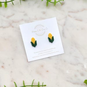 Könnte beinhalten: Ein Paar maisförmige Ohrstecker aus Polymer Clay. Die Ohrringe zeigen gelbe Maiskörner und grüne Blätter. Die Ohrringe werden auf einer weißen Karte mit dem Text "Happy Little Clay Co." präsentiert.