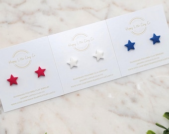 May include: Three pairs of star-shaped stud earrings in red, white, and blue. The earrings are presented on white cards with the text "Happy Little Clay Co" and "Handmade Polymer Clay Jewelry". The background is a marble surface with green foliage.