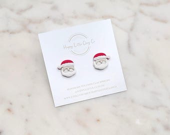 Puede incluir: Pendientes de botón de Papá Noel hechos de arcilla polimérica. Cada pendiente presenta una barba blanca, un gorro rojo y una cara sonriente. Los pendientes se muestran en una tarjeta blanca con el texto "Happy Little Clay Co."