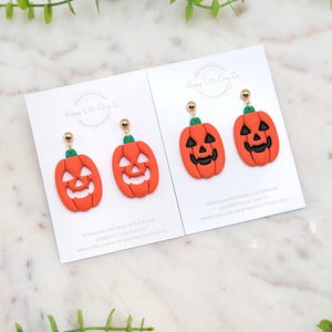 May include: Two pairs of orange pumpkin earrings with black carved faces and green stems, displayed on white cards. The earrings have gold-colored ball studs. The cards have the text "Happy Little Clay Co" and "HANDMADE POLYMER CLAY JEWELRY".