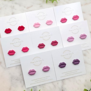Puede incluir: Surtido de pendientes de arcilla polimérica en varios tonos de rosa, rojo y morado. Cada par de pendientes tiene forma de labios. Los pendientes se muestran en tarjetas blancas con el texto "Happy Little Clay Co."