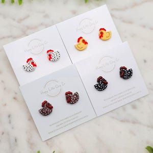 Könnte beinhalten: Vier Paar handgefertigte Polymer Clay Chicken Ohrringe. Jedes Paar hat eine andere Farbe und ein anderes Muster, darunter Schwarzweiß, Braunweiß, Gelbweiß und Rotweiß. Die Ohrringe werden auf weißen Karten mit dem Text "Happy Little Clay Co." präsentiert.
