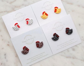 May include: Four pairs of handmade polymer clay chicken earrings. Each pair features a different colour and pattern, including black and white, brown and white, yellow and white, and red and white. The earrings are displayed on white cards with the text "Happy Little Clay Co."