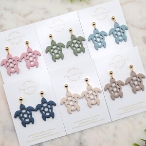 May include: Polymer clay turtle earrings in shades of pink, green, blue, and beige, displayed on white cards. Each pair has gold-coloured posts. The earrings are handmade and feature a cut-out design. The cards have the text "Happy Little Clay Co".
