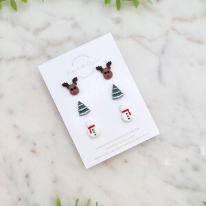 May include: A set of three handmade polymer clay earrings featuring a reindeer, a Christmas tree, and a snowman. The earrings are displayed on a white card with the text "Happy Little Clay Co."
