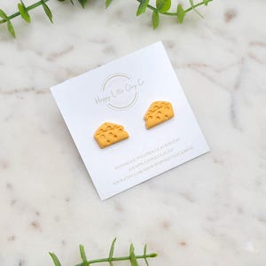 Op de afbeelding: Een paar gele polymer clay oorbellen in de vorm van plakjes Zwitserse kaas. De oorbellen zijn handgemaakt en worden verkocht door Happy Little Clay Co. op Etsy.
