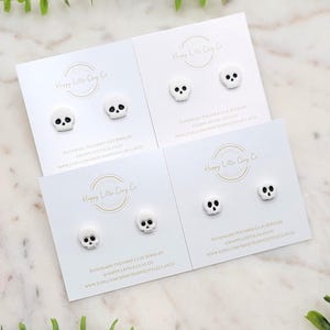 May include: Four pairs of white skull stud earrings on white cards. Each card has the text "Happy Little Clay Co" and "Handmade Polymer Clay Jewelry" with a website address. The earrings are a Halloween or gothic theme.