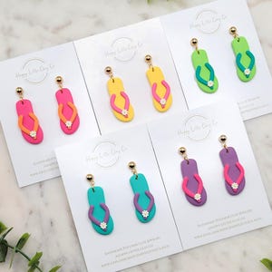 Op de afbeelding: Een verzameling kleurrijke polymeerklei oorbellen in de vorm van slippers. Elke paar heeft een andere kleurencombinatie, waaronder roze, geel, groen, turquoise en paars, met een kleine witte bloem. De oorbellen worden op witte kaarten getoond.