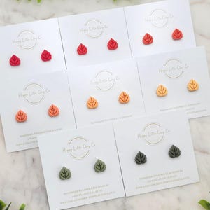 May include: A collection of leaf-shaped stud earrings in various colors, including red, orange, yellow, and green. Each pair is displayed on a white card with the text "Happy Little Clay Co" and "Handmade Polymer Clay Jewelry".