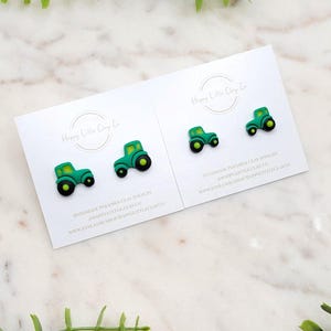 May include: Two pairs of green tractor-shaped polymer clay earrings on white cards. The tractors have black wheels and yellow accents. The cards have the text "Happy Little Clay Co" and "Handmade Polymer Clay Jewelry".
