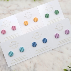 May include: Six pairs of handmade polymer clay earrings in various colors, including pink, yellow, green, blue, and purple. Each pair features a round stud with a striped pattern. The earrings are displayed on white cards with the brand name "Happy Little Clay Co."