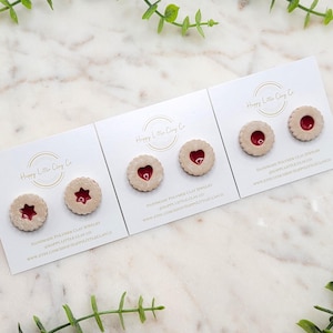 Puede incluir: Tres juegos de pendientes hechos a mano de arcilla polimérica. Cada juego presenta un diseño diferente: una estrella, un corazón y un círculo. Los pendientes son todos blancos con centros rojos. Las tarjetas en las que se muestran tienen el texto "Happy Little Clay Co." e información sobre la tienda.