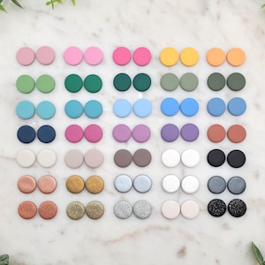 May include: Assortment of 20 pairs of round, colorful, and textured earrings. The earrings are arranged in rows on a white marble surface.